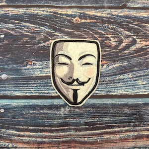 May include: Embroidered patch depicting the Guy Fawkes mask, with a white and grey design and black border. The mask has a smiling expression. The patch is set against a wooden background.
