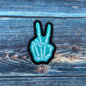 Puede incluir: Un parche de color azul turquesa en forma de signo de paz con un diseño de mano de esqueleto. El parche está bordeado de negro.