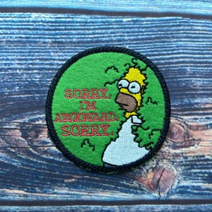 May include: A green embroidered patch featuring a yellow cartoon character peeking out from behind a bush. The text "Sorry, I'm awkward, Sorry." is embroidered in red on the patch.