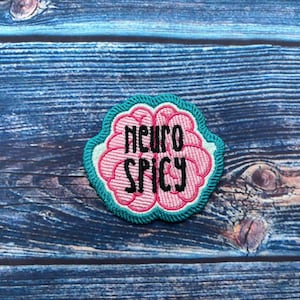 Può includere: Una toppa ricamata rosa e turchese a forma di cervello con il testo "neuro spicy" in nero.