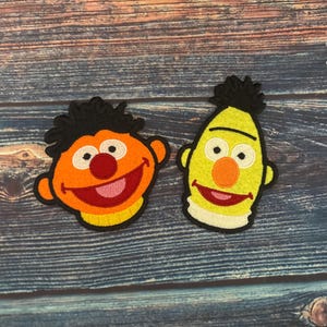 Op de afbeelding: Twee geborduurde patches met gezichten van stripfiguren. Eén is oranje met een rode neus en roze mond, de andere is geel met een oranje neus en roze mond. Beide hebben zwart haar en lachen.