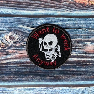 Peut inclure: Écusson rond noir avec un motif de squelette blanc et le texte "Went to work anyway" en rouge et blanc. Le patch est circulaire et a une bordure noire. Le fond est une surface en bois bleu et marron.