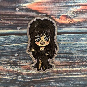 Pode incluir: Patch bordado com uma personagem de desenho animado com cabelo preto e um vestido preto. A personagem tem olhos azuis e uma expressão amigável. O patch é delineado em cinza e está sobre um fundo de grão de madeira.