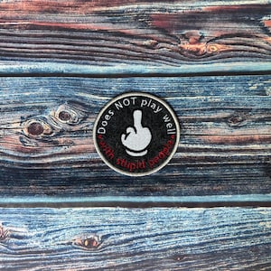 Peut inclure: Écusson rond brodé avec le texte "Does NOT play well with stupid people". Le patch a une bordure noire et une main blanche faisant un geste obscène. Le patch est sur une surface en bois bleu et marron.