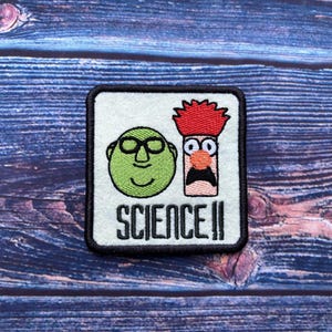 Peut inclure: Écusson carré avec deux personnages de dessins animés, l'un vert avec des lunettes et l'autre avec des cheveux roux et une expression surprise. Le mot "SCIENCE!!" est imprimé en noir en dessous. L'écusson a une bordure noire et est posé sur un fond en bois bleu.