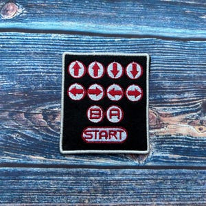 May include: Black and white embroidered patch with red directional arrows, a B and A button, and a START button. The patch is on a blue wooden surface.
