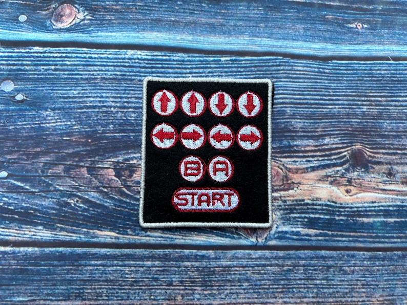 Peut inclure: Patch brod&eacute; noir et blanc avec des fl&egrave;ches directionnelles rouges, un bouton B et A, et un bouton START. Le patch est sur une surface en bois bleu.