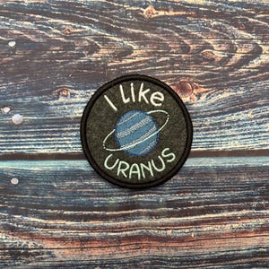 May include: A round, embroidered patch with the text "I like Uranus" in white. The patch features a blue planet with a ring. The patch is on a dark gray background with a black border.