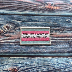 Pode incluir: Remendo bordado com o texto "FUCK YOU, I WON'T BE WHAT YOU TELL ME." O remendo apresenta um design com listras rosa, brancas e verde-azuis. O remendo é retangular com uma borda bege clara.