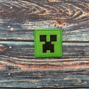 Op de afbeelding: Een vierkant, groen embleem met een zwart gepixeld Creeper-gezicht uit de populaire game Minecraft. De patch is waarschijnlijk een opstrijk- of opnaai-applicatie, perfect voor het aanpassen van kleding of accessoires. Op een houten achtergrond.
