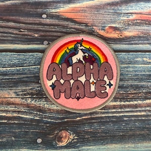 May include: Circular patch featuring the text "ALPHA MALE" in a bubble font, a rainbow, and a unicorn design. The patch has a pink backdrop with a grey border, set against a wood grain background.