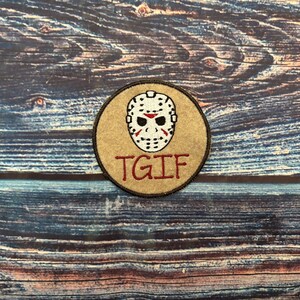 May include: A circular patch with a white hockey mask with red details on a tan background. The red text "TGIF" is below the mask. The patch has a brown border and is on a wood grain surface.