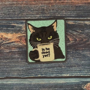 以下が含まれることがあります： 黄色い目をした黒猫がマグカップを持っている刺繍パッチ。マグカップには「Is he dead yet?」と書かれています。パッチはターコイズブルーの背景とグレーの縁取りがあります。パッチは正方形です。