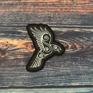 May include: Black patch with a white embroidered raven design. The raven is depicted in flight, with detailed wing feathers and swirling patterns. The patch is set against a dark wooden background.
