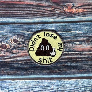 May include: A circular patch with the words "Didn't lose my shit" and a cartoon poop emoji with a middle finger. The patch is yellow with black and white details. It rests on a blue and brown wooden surface.