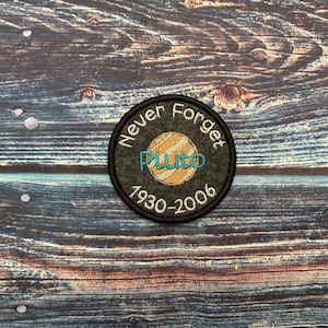 Op de afbeelding: Een ronde, geborduurde patch met de tekst "Never Forget Pluto" in het wit, gebogen aan de bovenkant. Het midden toont een gestileerde Pluto met de tekst "Pluto" in turquoise. De onderkant leest "1930-2006".