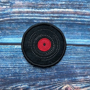 May include: A black vinyl record with a red centre label, embroidered on a fabric patch. The patch is on a blue wooden background.