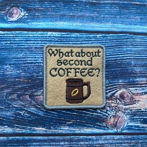 Puede incluir: Parche bordado con el texto "What about second COFFEE?" encima de una taza de café marrón. El parche tiene un fondo beige con un borde azul y está colocado sobre un fondo de madera azul.