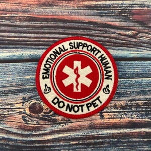 May include: A white and red round embroidered patch with the text "Emotional Support Human Do Not Pet" and a red medical symbol with a snake inside.