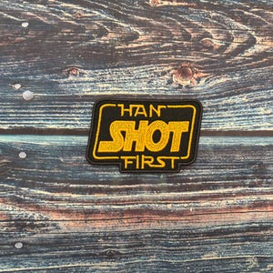 May include: A black and yellow patch with the text "HAN SHOT FIRST" in a stylized font. The patch has a black border and is set against a weathered wood background.