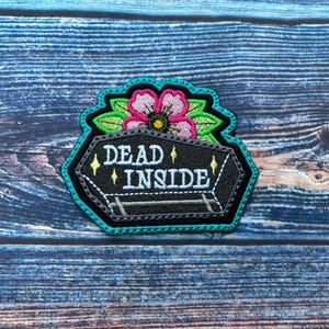 Könnte beinhalten: Schwarzer und weißer gestickter Aufnäher mit einem Sarg und einer Blume. Der Sarg trägt den Text "DEAD INSIDE".