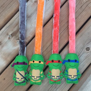 Peut inclure: Quatre sucettes glacées congelées dans des emballages en plastique transparent avec du jus congelé coloré à l'intérieur. Chaque sucette est décorée d'un personnage de la Tortue Ninja Adolescente en feutre.