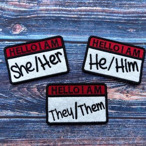 Può includere: Tre etichette per nomi in feltro bianco con bordi rossi e testo nero. Il testo sulle etichette recita "HELLO I AM She/Her", "HELLO I AM He/Him" e "HELLO I AM They/Them".