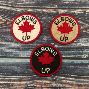 May include: Three embroidered patches with a red maple leaf and the text "Elbows Up". The patches are on a wooden background. The patches are white, beige, and black.