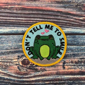 Op de afbeelding: Een ronde, blauwe en gele geborduurde patch met een groene kikker en de tekst "Vertel me niet om te lachen".