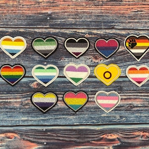 Puede incluir: Un conjunto de parches en forma de corazón con varios diseños de banderas del orgullo LGBTQ+. Cada parche presenta un borde negro y una combinación de colores única que representa diferentes identidades. Los parches están dispuestos sobre una superficie de madera.