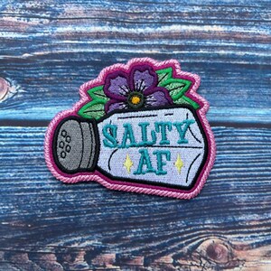 May include: A white salt shaker with a pink border and a purple flower on top. The shaker has the text "Salty AF" in blue embroidery.