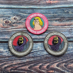 Könnte beinhalten: Drei gestickte Aufnäher mit Zeichentrickfiguren. Ein Aufnäher hat einen rosa Rand mit dem Text "I SAID HEY WHAT'S GOING ON!" und eine blonde Figur. Zwei graue Aufnäher zeigen eine Figur mit Totenkopf und Text.