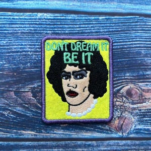 May include: A yellow and purple embroidered patch with a cartoon image of a person with dark hair and the text "Don't Dream It Be It".
