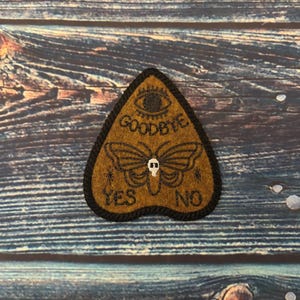May include: A brown felt ouija board patch with black stitching. The board features a black and white butterfly with a skull, a black eye, and the words "Goodbye", "Yes", and "No".