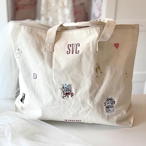 May include: Cream-colored tote bag with embroidered details. Features the initials "SVC" in burgundy, along with small illustrations of a ring, flowers, champagne flutes, a heart, and a cake. The date IX.XXVII.XXV is also embroidered.