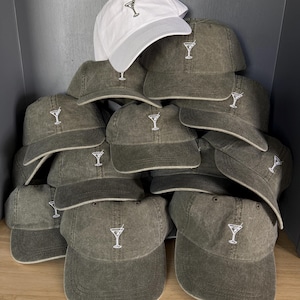 May include: A collection of baseball caps stacked together. One white cap sits on top of several olive green caps. Each cap features a small, white embroidered martini glass icon centred on the front panel above the curved brim.
