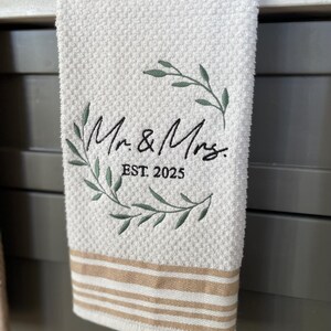 Op de afbeelding: Witte keukendoek met bruine strepen, met een geborduurde krans met groene bladeren en de tekst "Mr. & Mrs. EST. 2025".