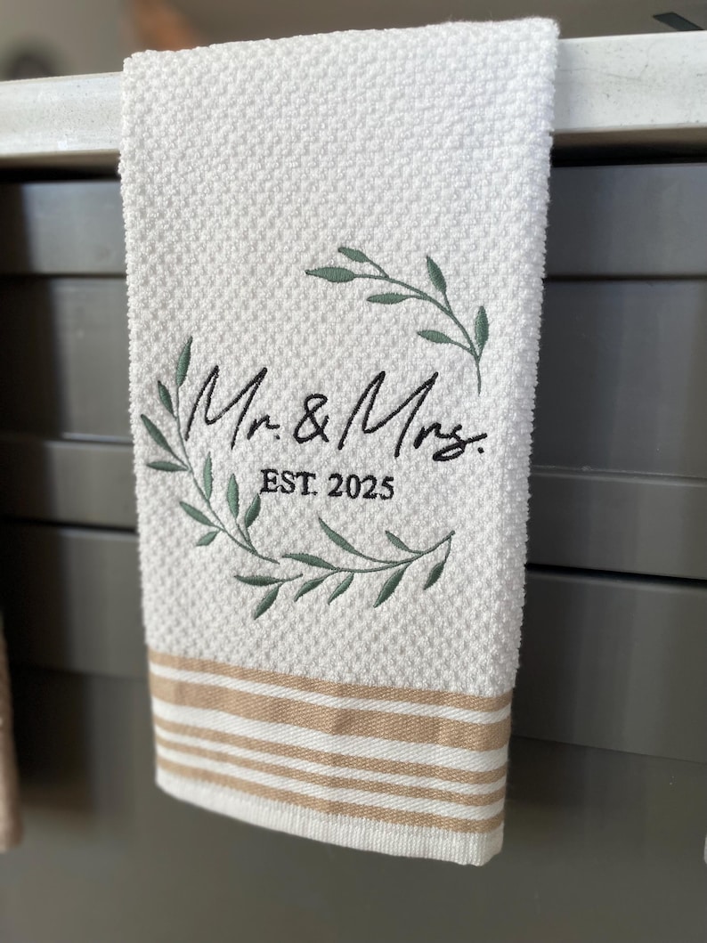 Op de afbeelding: Witte keukendoek met bruine strepen, met een geborduurde krans met groene bladeren en de tekst "Mr. & Mrs. EST. 2025".