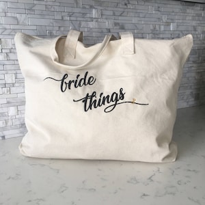 May include: A white canvas tote bag with black embroidered text that says "bride things".