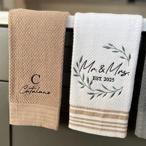 Op de afbeelding: Twee keukendoeken hangen aan een rek. E&eacute;n is beige met de tekst "C Catalano" geborduurd erop. De andere is wit met een groene krans en de tekst "Mr. & Mrs. EST 2025" geborduurd erop.
