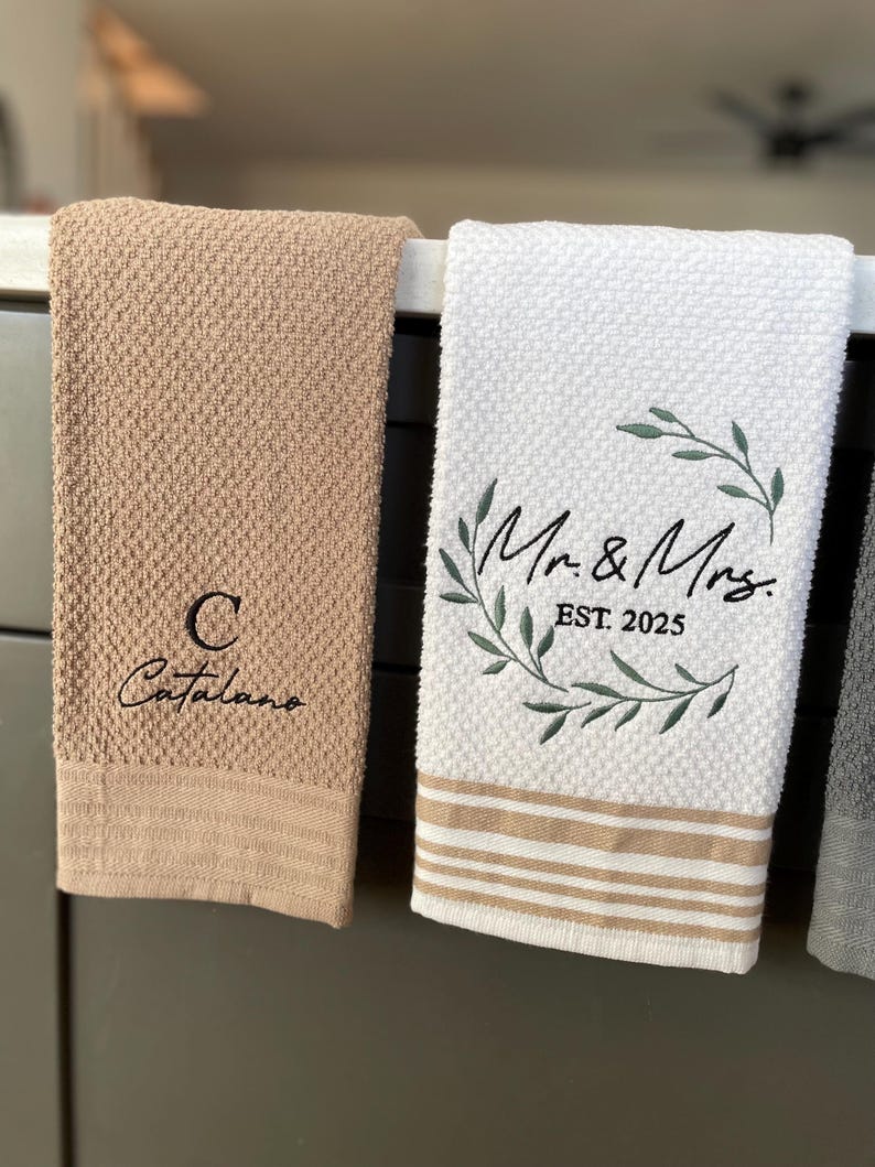 Op de afbeelding: Twee keukendoeken hangen aan een rek. E&eacute;n is beige met de tekst "C Catalano" geborduurd erop. De andere is wit met een groene krans en de tekst "Mr. & Mrs. EST 2025" geborduurd erop.