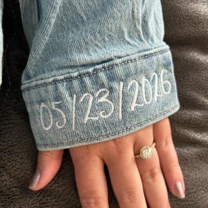 May include: Light blue denim jacket cuff with embroidered date "05/23/2026" in white thread. A gold ring with a round, diamond-studded halo is on a hand with manicured nails.