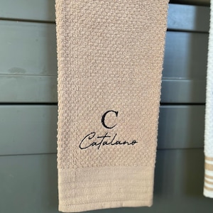 Op de afbeelding: Een beige keukendoek met een zwarte geborduurde letter "C" en de naam "Catalano" in cursief.
