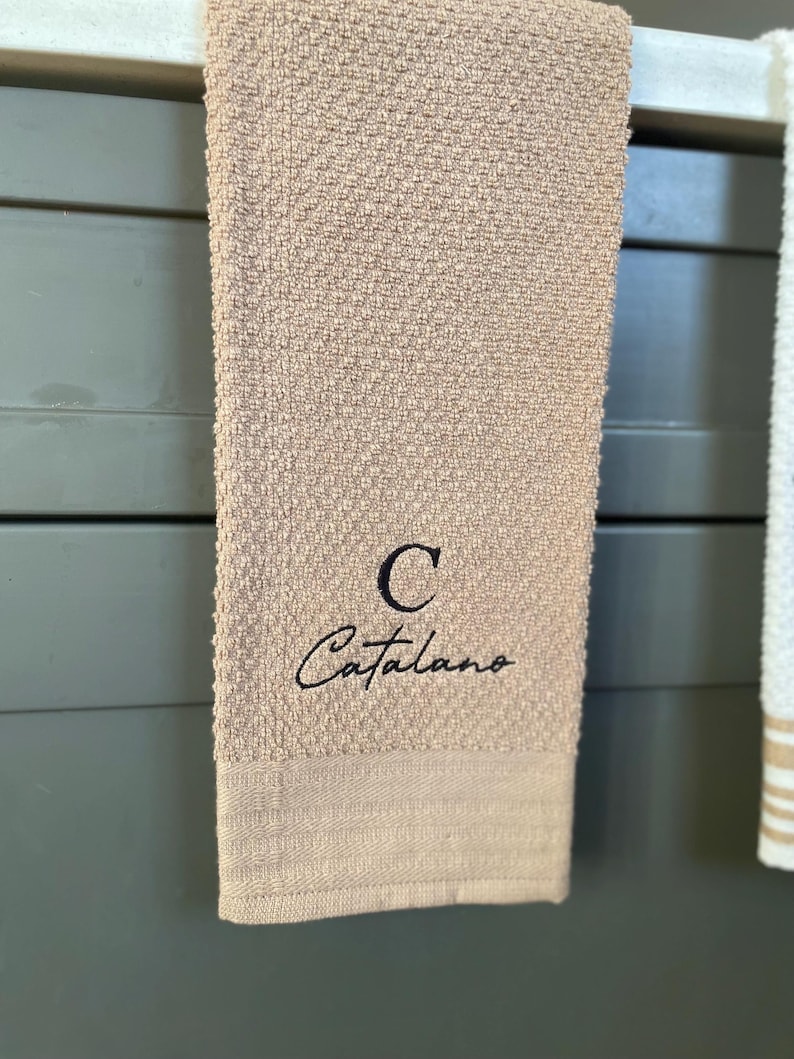 Op de afbeelding: Een beige keukendoek met een zwarte geborduurde letter "C" en de naam "Catalano" in cursief.