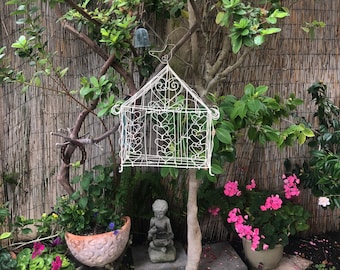 May include: A white metal birdcage with a heart design hangs from a tree branch in a garden setting. A small gray stone statue of a person sits on the ground in front of the cage. Pink flowers bloom in a pot to the right of the cage.