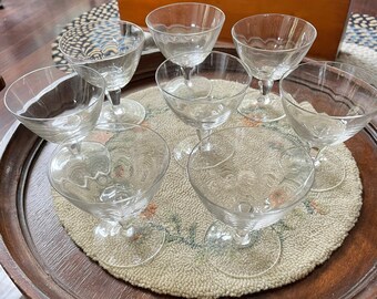 May include: A set of eight clear glass stemmed drinking glasses on a wooden table with a floral patterned rug.