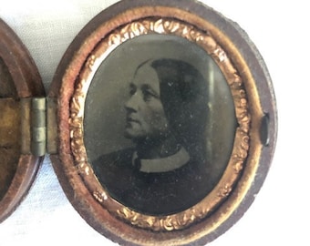 May include: A small, round, wooden locket with a gold-colored metal frame. The locket is open, revealing a black and white photograph of a man with a beard.