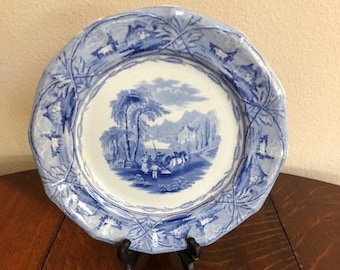 May include: A blue and white porcelain plate with a decorative border and a scene of a rural landscape with figures and animals. The plate is on a wooden stand.