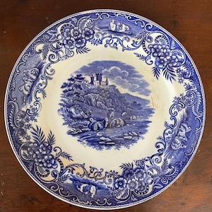 May include: A round, antique blue and white ceramic plate. The plate features a central landscape scene with a castle, figures, and animals. The border is decorated with floral and scroll patterns. The plate is set on a dark wooden surface.