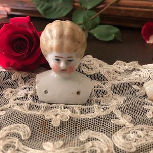 May include: A porcelain doll head with blonde hair and a painted face. The head is on a white lace doily.